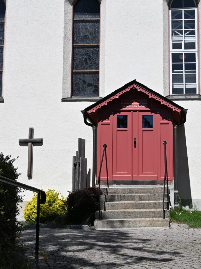 Rote Tür mit Treppen, neben einer weißen Wand und einem Kreuz, umgeben von Pflanzen.