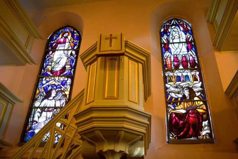 Buntglasfenster in einer Kirche, neben einer Holzkanzel mit Kreuz.